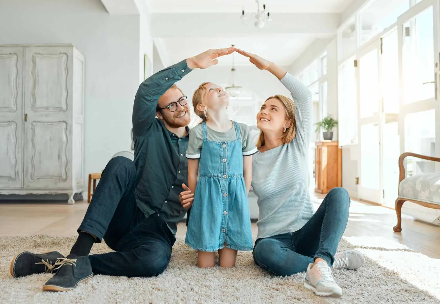 A family sitting on the living room floor with the parents making a roof shape over their daughter